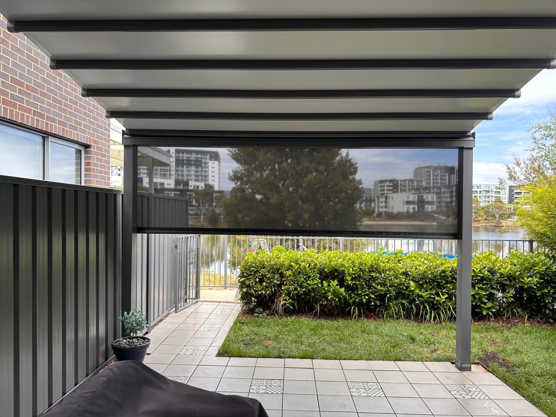 Helioscreen All Seasons retractable roof in closed position with HMX 100 external drop screen deployed, installed at a residential property near the Greenway, Canberra ACT, anthracite aluminium frame, garden and waterway in background